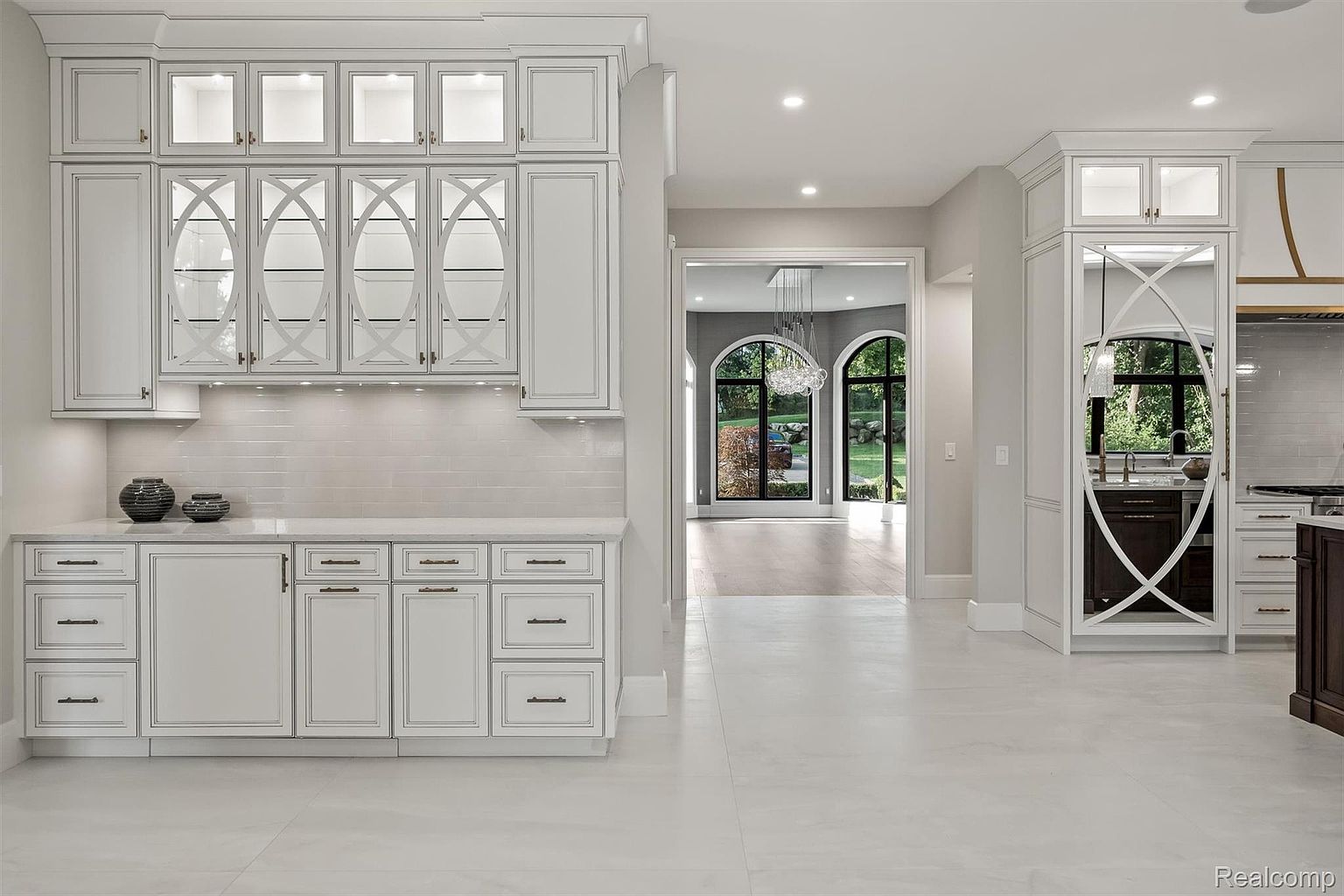 This is a bright and spacious kitchen featuring custom white cabinetry with glass-fronted upper cabinets and detailed trim. The kitchen is open to a hallway with large arched windows providing natural light and views of the outdoors. The overall impression is one of luxury and sophistication, perfect for a high-end real estate listing.
