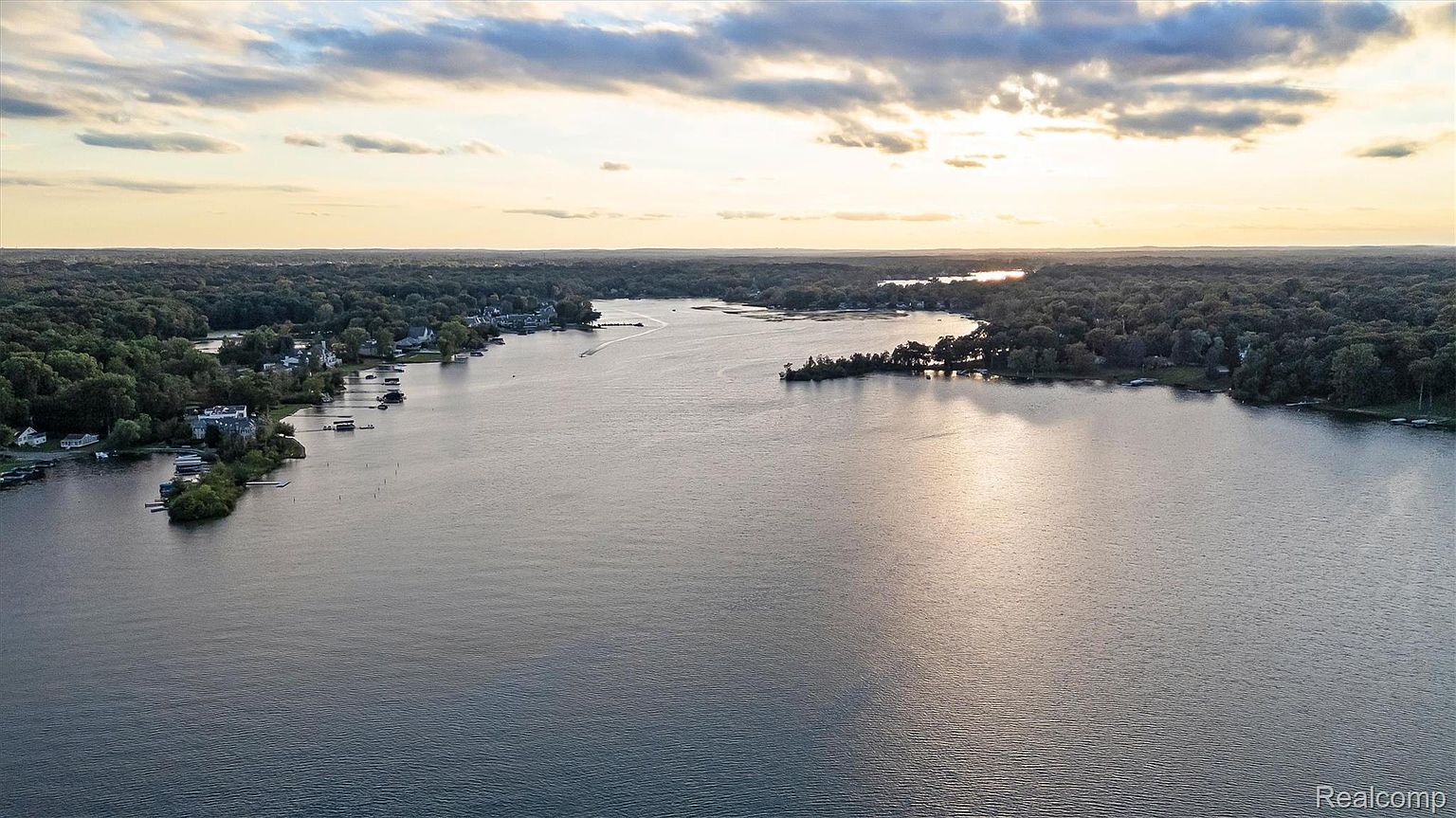 This aerial view showcases a serene lake surrounded by lush, dense trees and residential properties along the shoreline. The water reflects the soft light of the setting sun, creating a tranquil and inviting atmosphere. The landscape suggests a peaceful and secluded living environment, perfect for those seeking a retreat from city life.