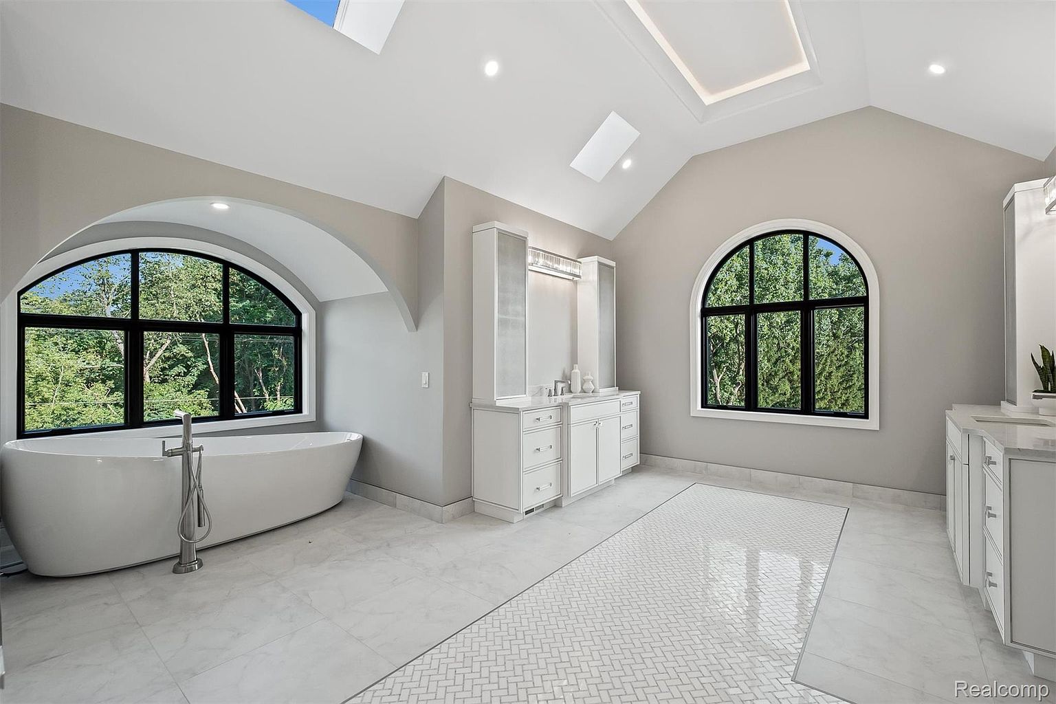 This is a luxurious primary bathroom featuring a freestanding soaking tub positioned beneath a large arched window, offering a view of lush greenery. The space is adorned with white marble flooring, a herringbone-patterned rug, and elegant white cabinetry with ample storage. Skylights and recessed lighting illuminate the room, creating a bright and airy atmosphere.