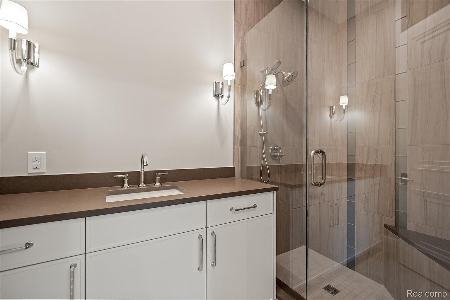 This is a primary bathroom featuring a modern design with a brown countertop and white cabinets. The shower is enclosed with glass and has a built-in bench. The walls are tiled in a neutral tone, and the lighting is provided by sconces.