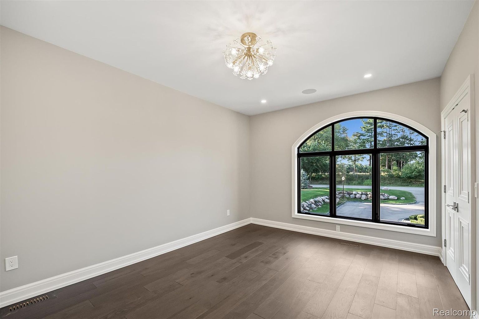 This is an interior shot of a bedroom featuring dark hardwood floors, light gray walls, and white trim. A large arched window with black frames provides natural light and a view of the outdoors. A decorative chandelier hangs from the ceiling, adding a touch of elegance to the room.