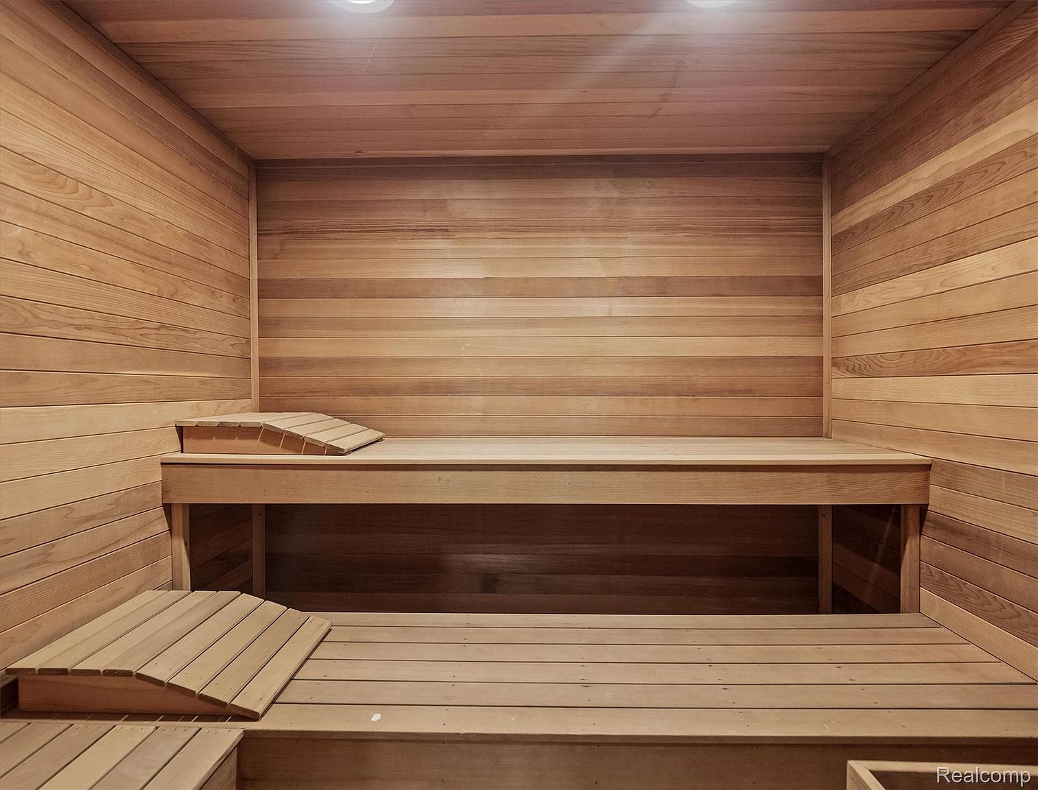 This is an interior shot of a sauna, featuring wooden walls, benches, and flooring. The sauna is well-lit with overhead lighting, creating a warm and inviting atmosphere. The benches are arranged for comfortable seating, and the overall design is clean and minimalist.