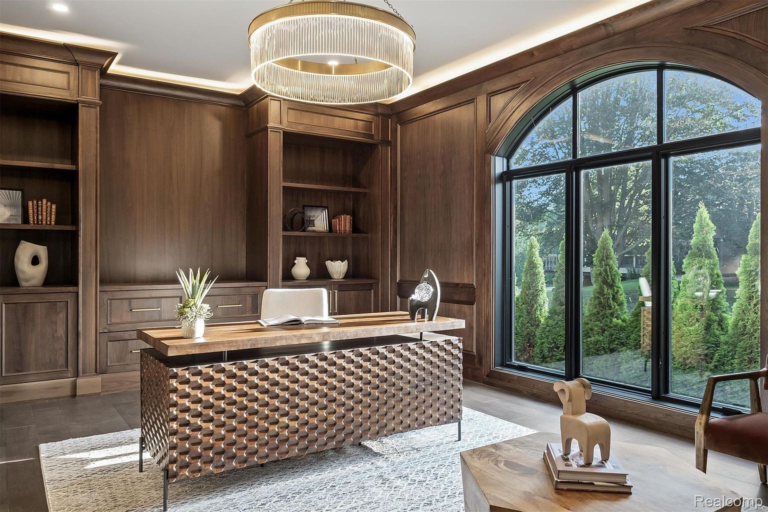 This is an interior shot of a luxurious home office featuring rich wood paneling and built-in bookshelves. A unique, textured desk sits in the center of the room, complemented by a modern chandelier. A large arched window provides natural light and a view of the exterior greenery, enhancing the room's sophisticated and inviting atmosphere.