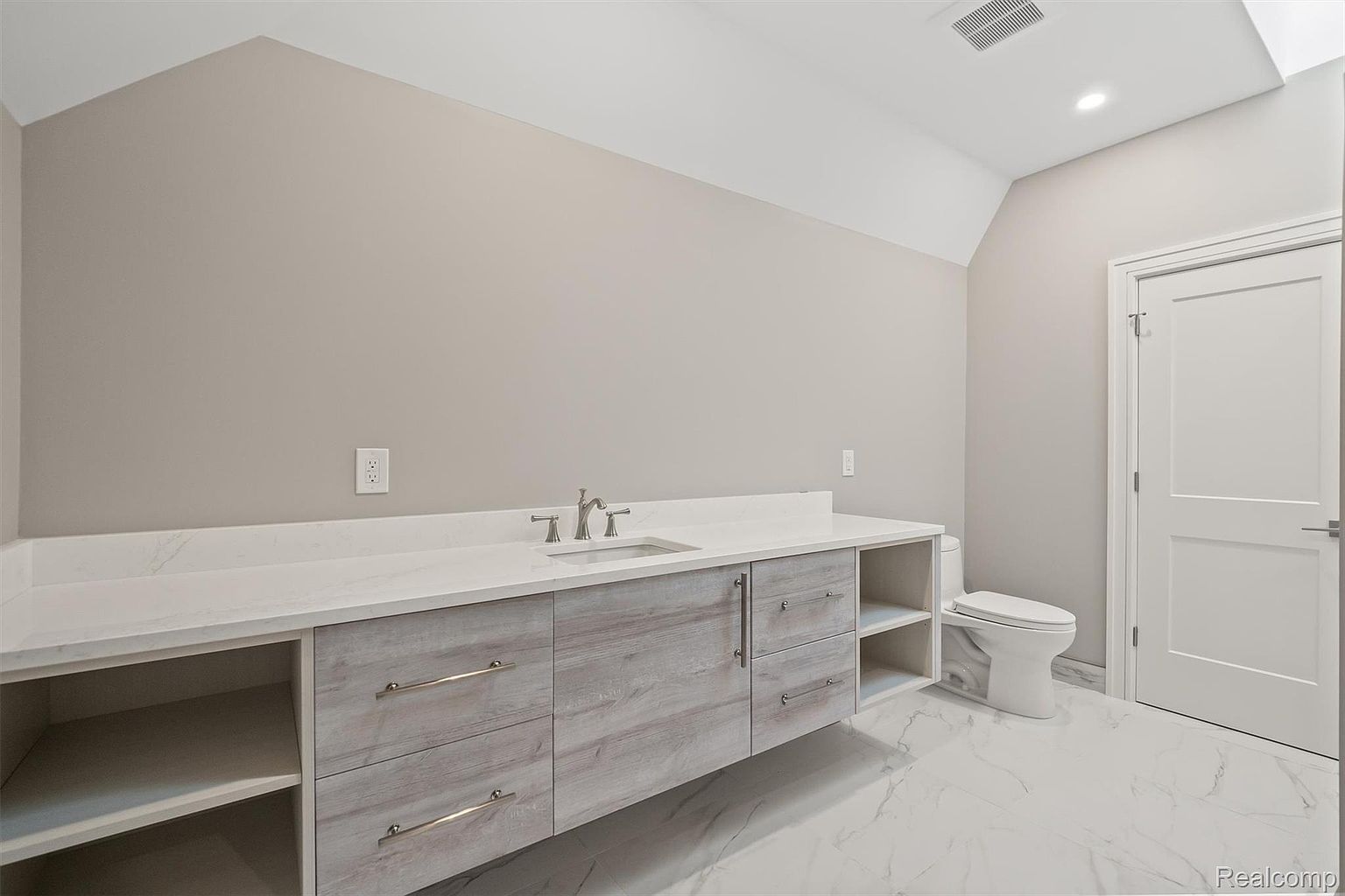 This is a bright and clean bathroom featuring a long vanity with a white countertop and light wood-grain cabinets. A modern sink and faucet are installed, and a toilet is positioned next to the vanity. The floor is white marble, and a white door is visible on the right side of the image.