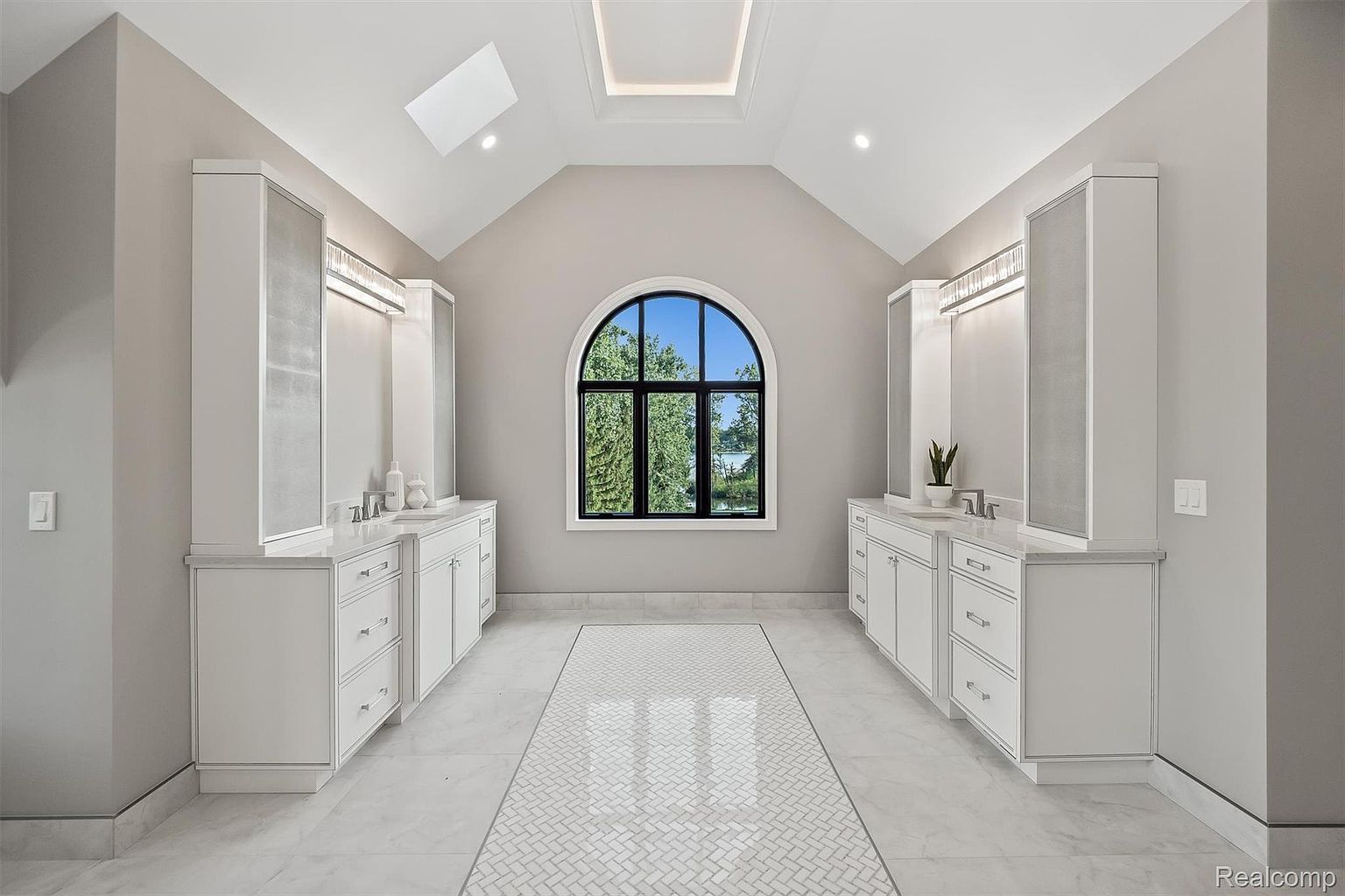 This is a spacious primary bathroom featuring dual white vanities with ample storage and countertop space. A large arched window provides natural light and a view of the outdoors. The flooring is a combination of large format tiles and a decorative herringbone pattern, adding visual interest to the room.