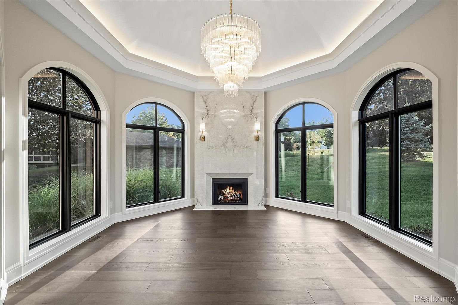 This is an interior shot of a sunroom or living room featuring a fireplace and large arched windows that provide ample natural light. The room is decorated with a large chandelier and has hardwood floors, creating an elegant and inviting atmosphere. The fireplace is surrounded by marble and flanked by sconces.