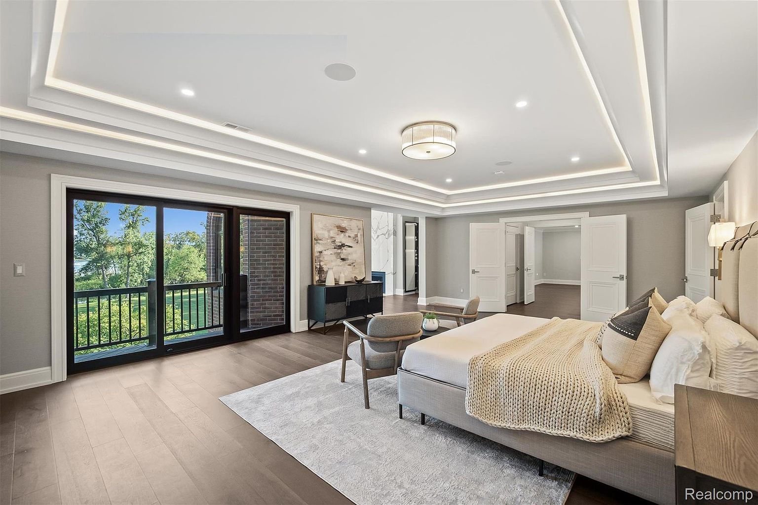 This is a spacious primary bedroom featuring hardwood floors, a tray ceiling with recessed lighting, and a large sliding glass door leading to a balcony. The room is furnished with a king-size bed, a sitting area with two chairs, and a dark wood dresser with artwork above it. The overall style is modern and luxurious.