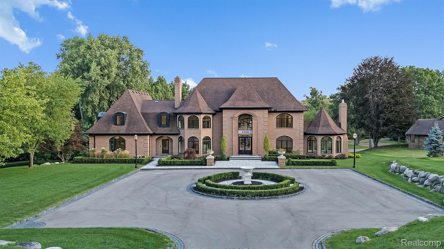 This is a grand front view of a large, luxurious brick home. The house features a circular driveway with a central fountain, manicured landscaping, and multiple arched windows. The overall impression is one of elegance and stately charm, perfect for a high-end real estate listing.