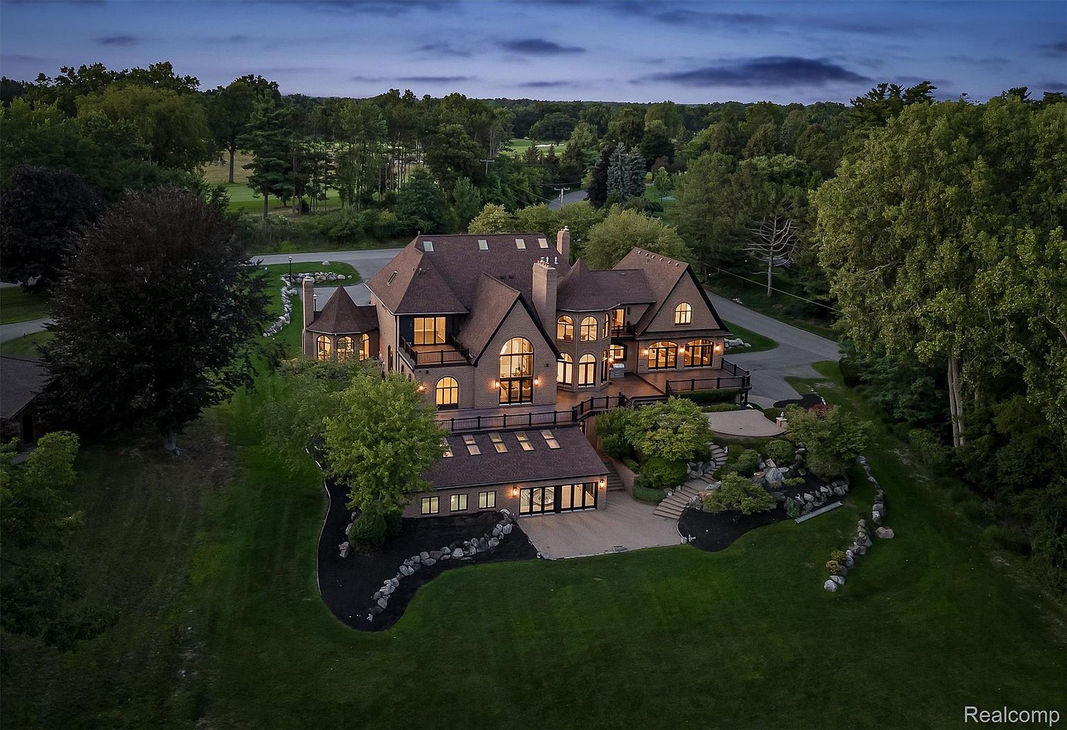 This aerial view showcases a grand, multi-story home with a brick exterior and a dark roof, surrounded by lush greenery and mature trees. The property features a large deck, a landscaped yard with stone accents, and a lower-level patio area. The overall impression is one of luxury and privacy, set within a serene, park-like setting.