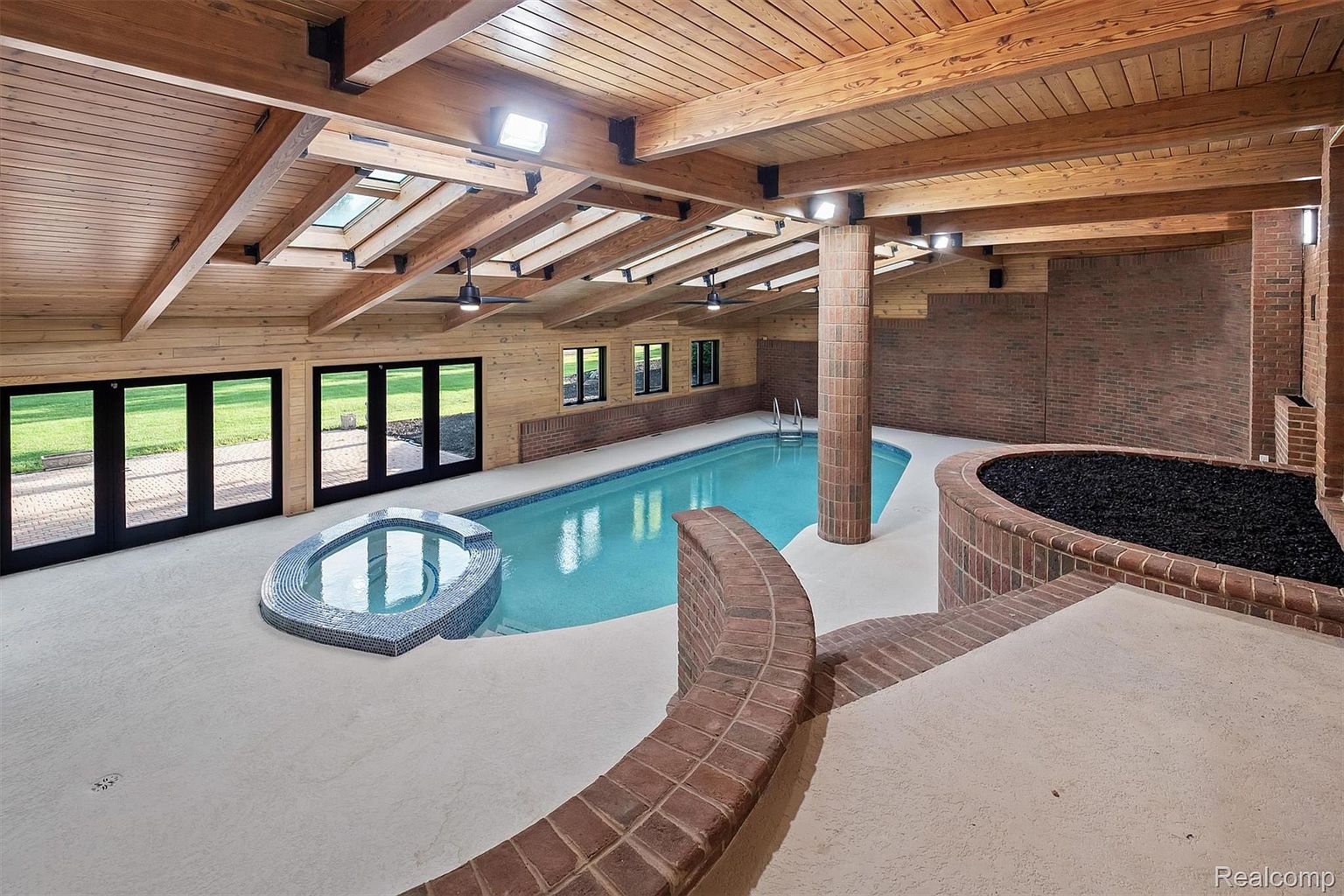 This image showcases an indoor pool and spa area, featuring a unique design with a brick accent wall and a wooden ceiling with skylights. The pool is surrounded by a concrete deck, and a raised brick planter adds a touch of greenery. The space is well-lit and appears to be part of a luxurious home.