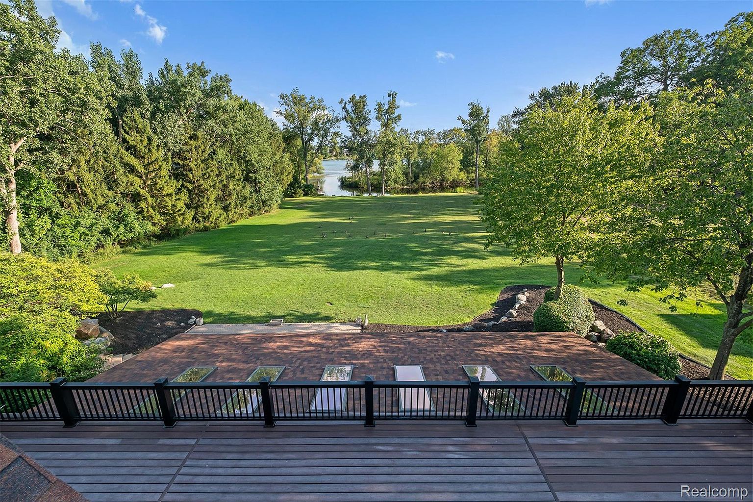 This exterior shot showcases a sprawling backyard with a lush green lawn extending towards a body of water, likely a lake or pond. A multi-level deck with dark railings provides an elevated view of the landscape, featuring skylights built into the lower level roof. The scene is framed by mature trees, creating a serene and private setting.