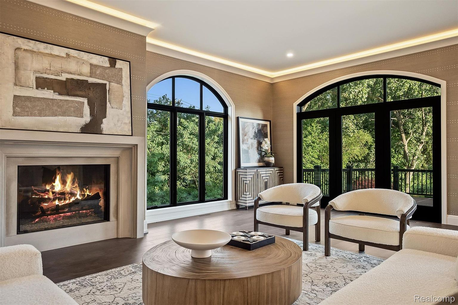 This is a beautifully staged living room featuring a fireplace with a large abstract painting above it. The room has two arched windows offering views of lush greenery, and two armchairs are placed around a round wooden coffee table. The overall aesthetic is elegant and inviting, perfect for showcasing a comfortable and stylish living space.