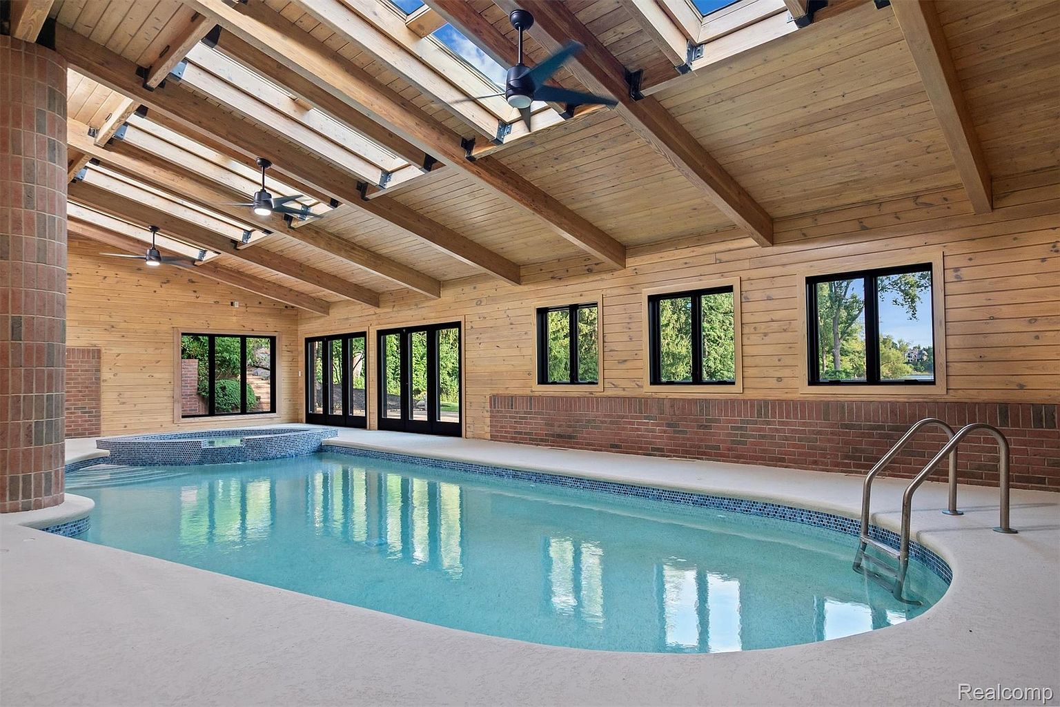 This image showcases an indoor pool and spa area, featuring a long rectangular pool with light blue water and a connected spa section. The room is enclosed with wood-paneled walls, brick accents, and multiple windows and doors that provide natural light. The ceiling has exposed wooden beams and skylights, enhancing the bright and airy atmosphere.