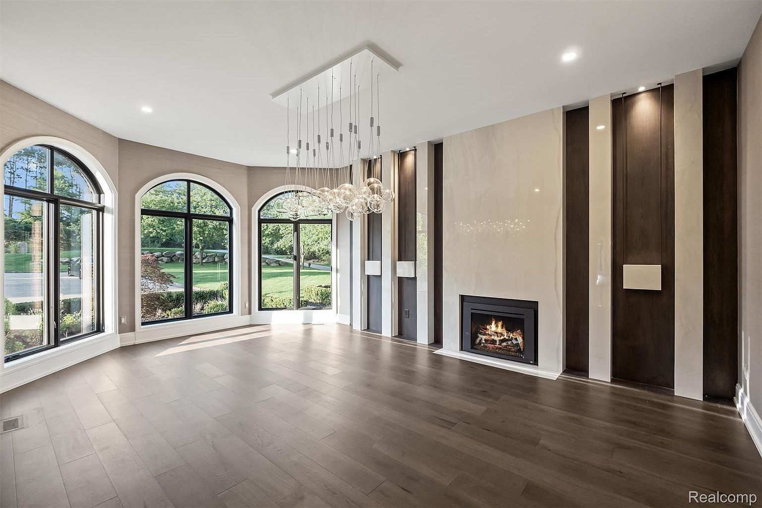 This is a spacious living room featuring hardwood floors, large arched windows offering natural light, and a modern fireplace with decorative paneling. A contemporary chandelier hangs from the ceiling, adding a touch of elegance. The room is empty, showcasing its size and potential for customization.