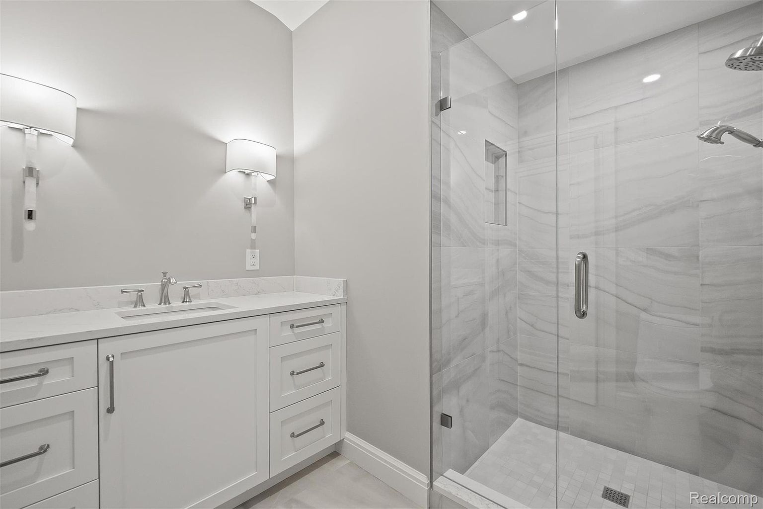 This is a well-lit primary bathroom featuring a white vanity with a marble countertop and modern fixtures. The room also includes a glass-enclosed shower with marble tiling and a rainfall showerhead. The overall impression is clean, bright, and luxurious, suggesting a high-end renovation or new construction.
