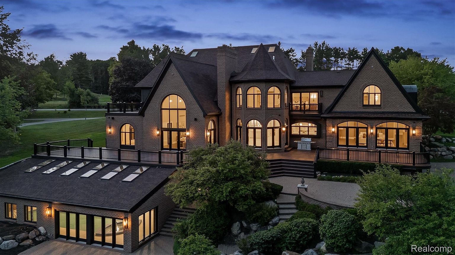 This is a rear exterior view of a large, luxurious brick home at dusk. The house features multiple arched windows, a multi-level deck, and a connected garage with skylights. The landscaping includes mature trees, shrubs, and a stone pathway, creating a serene and private backyard setting.