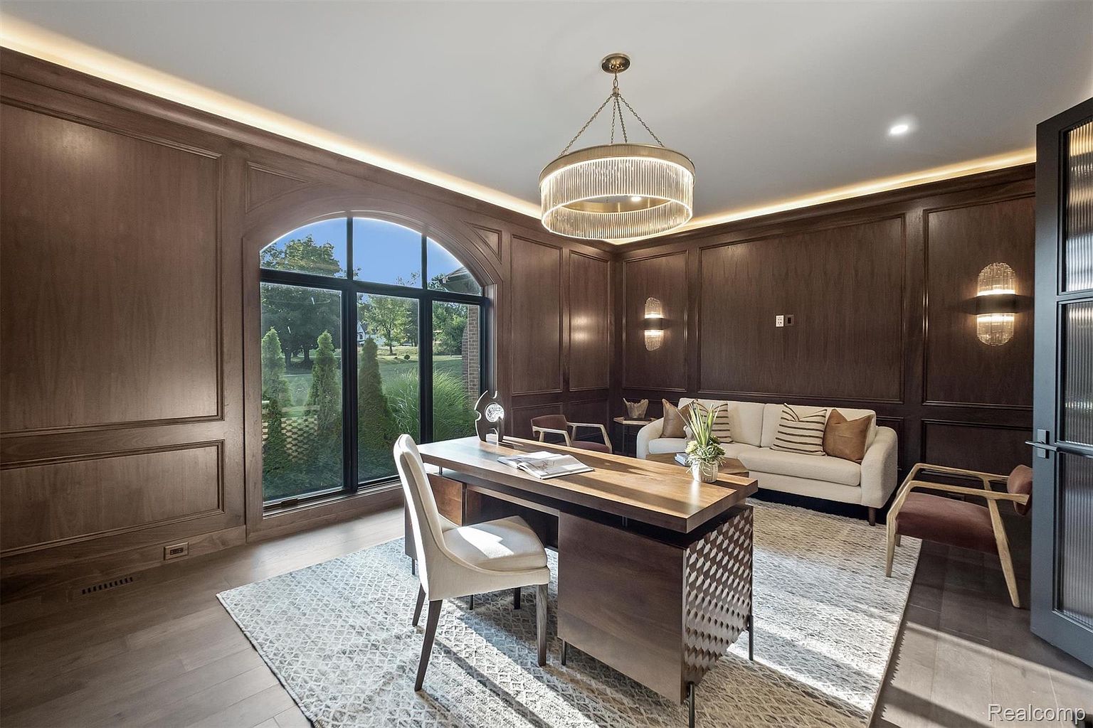 This is an interior shot of a luxurious home office. The room features dark wood paneling, a large arched window providing natural light, and a modern chandelier. A wooden desk and comfortable chair are centrally located, with a sofa and additional seating in the background, creating a sophisticated and inviting workspace.