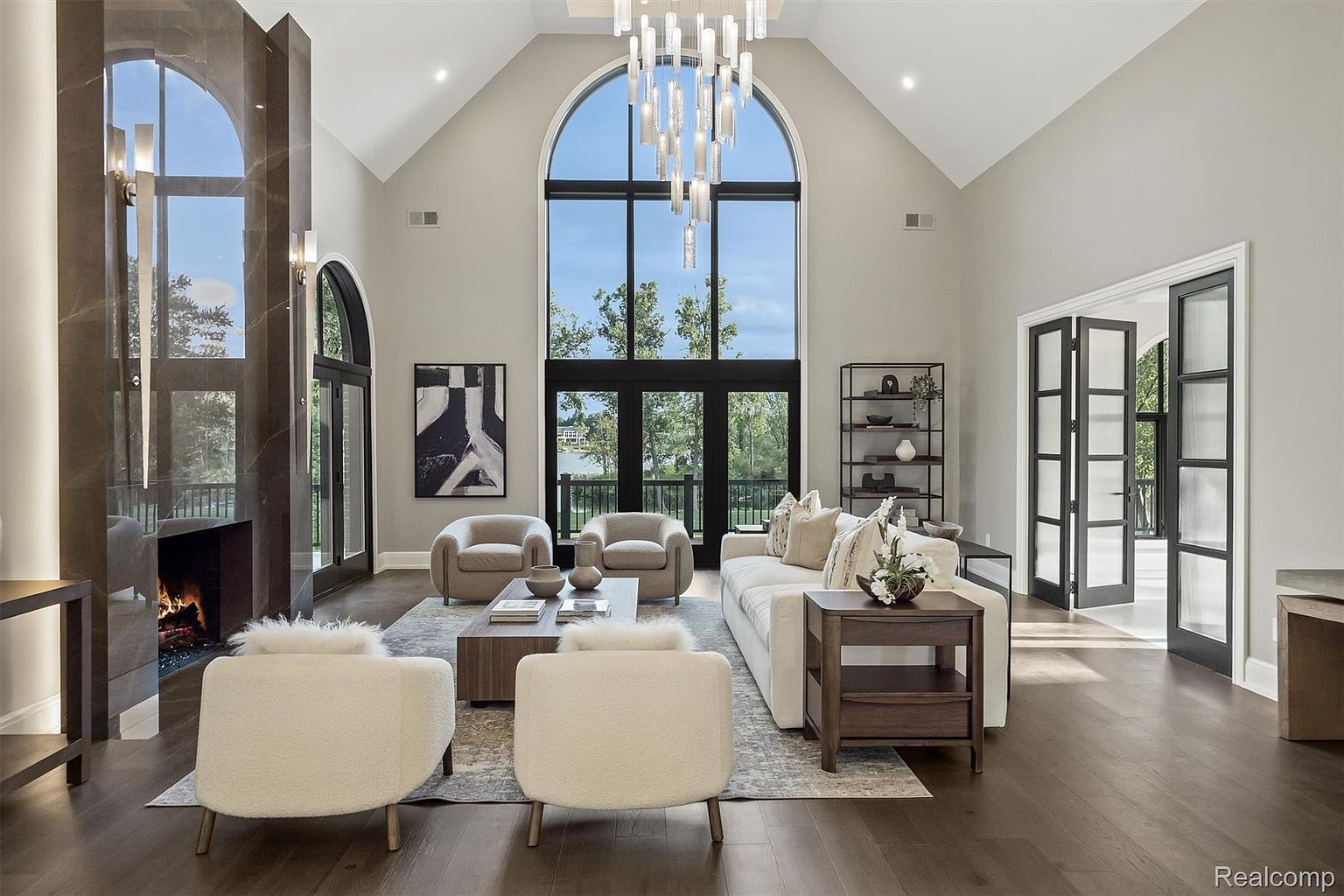 This is a spacious living room featuring a high vaulted ceiling and a large arched window that provides ample natural light. The room is furnished with a neutral-toned sofa, armchairs, and a coffee table, creating a comfortable and inviting atmosphere. A modern fireplace and a decorative chandelier add to the room's elegance and style.