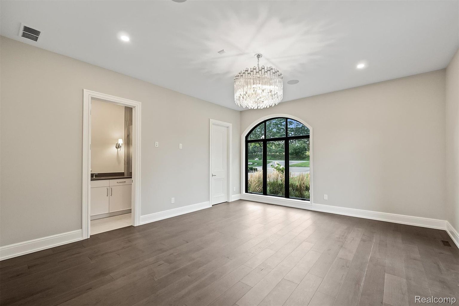 This is a spacious primary bedroom featuring dark hardwood floors and light beige walls. A large arched window provides natural light and a view of the outdoors. The room is illuminated by a modern chandelier, and there is an ensuite bathroom visible through a doorway.