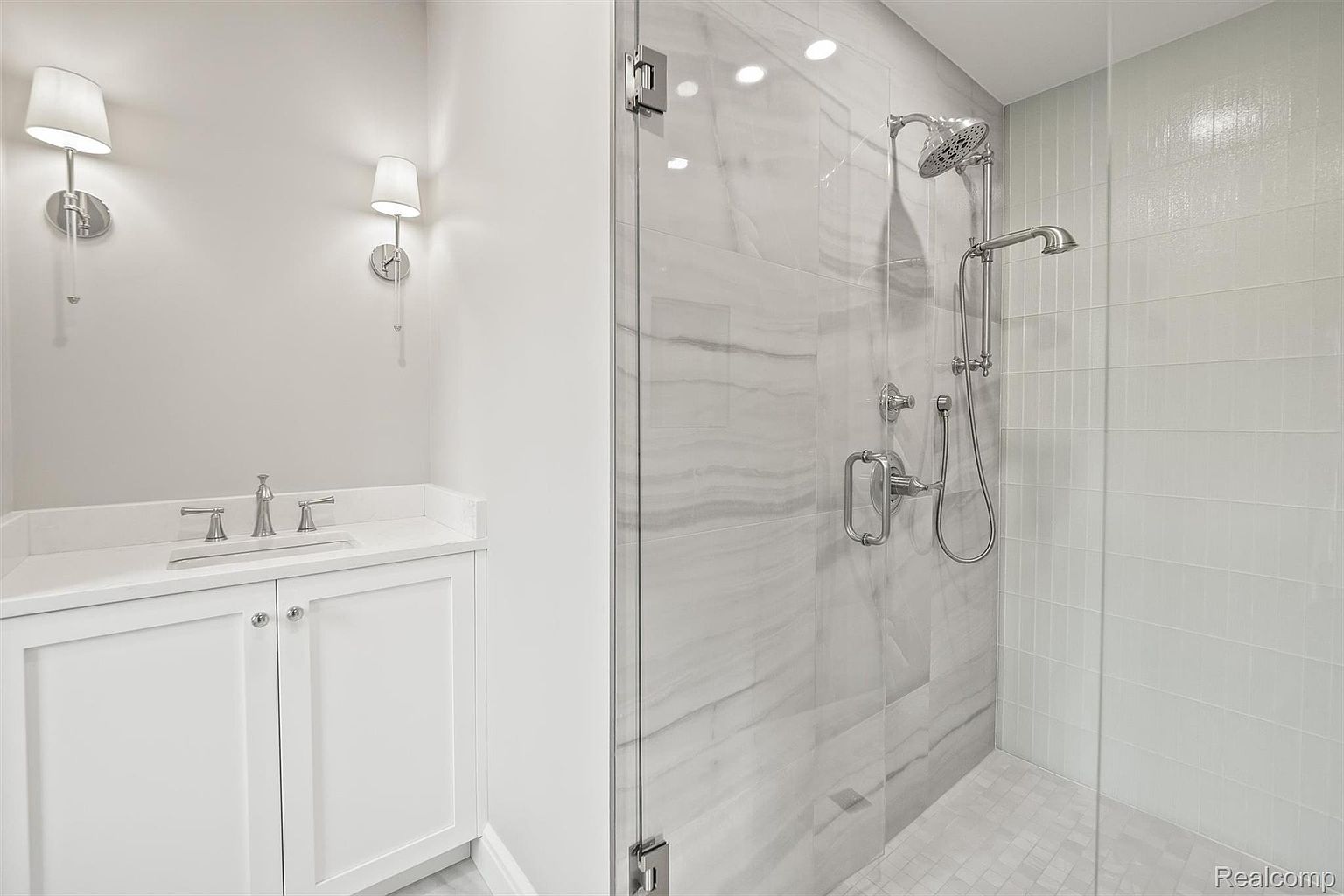 This is a well-lit bathroom featuring a modern design with a white vanity and countertop, complemented by two sconce lights. The shower area is enclosed with glass and showcases marble-like tiles and a contemporary shower system. The overall impression is clean, bright, and stylish, suggesting a recently renovated or well-maintained space.