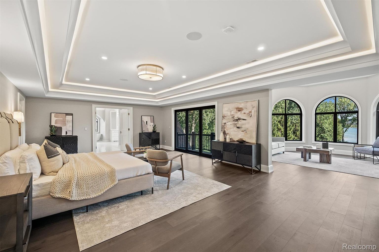 This is a spacious primary bedroom featuring a tray ceiling with recessed lighting, hardwood floors, and a neutral color palette. The room includes a large bed, seating area, and access to a balcony through black-framed sliding doors. Large arched windows offer views of the outdoors, creating a serene and luxurious atmosphere.