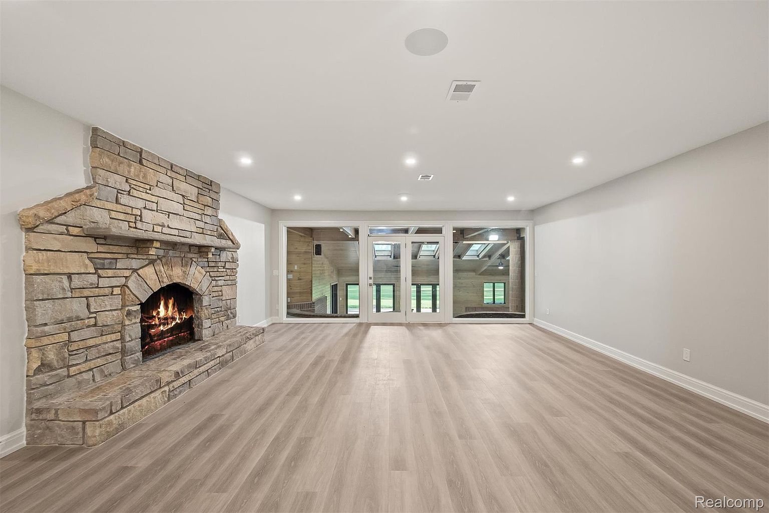This is an interior shot of a living room featuring a stone fireplace on the left side of the frame. The room has light-colored wood flooring and white walls, creating a bright and spacious feel. Large windows or glass doors are visible at the back, offering a view to the outside and allowing natural light to flood the room.