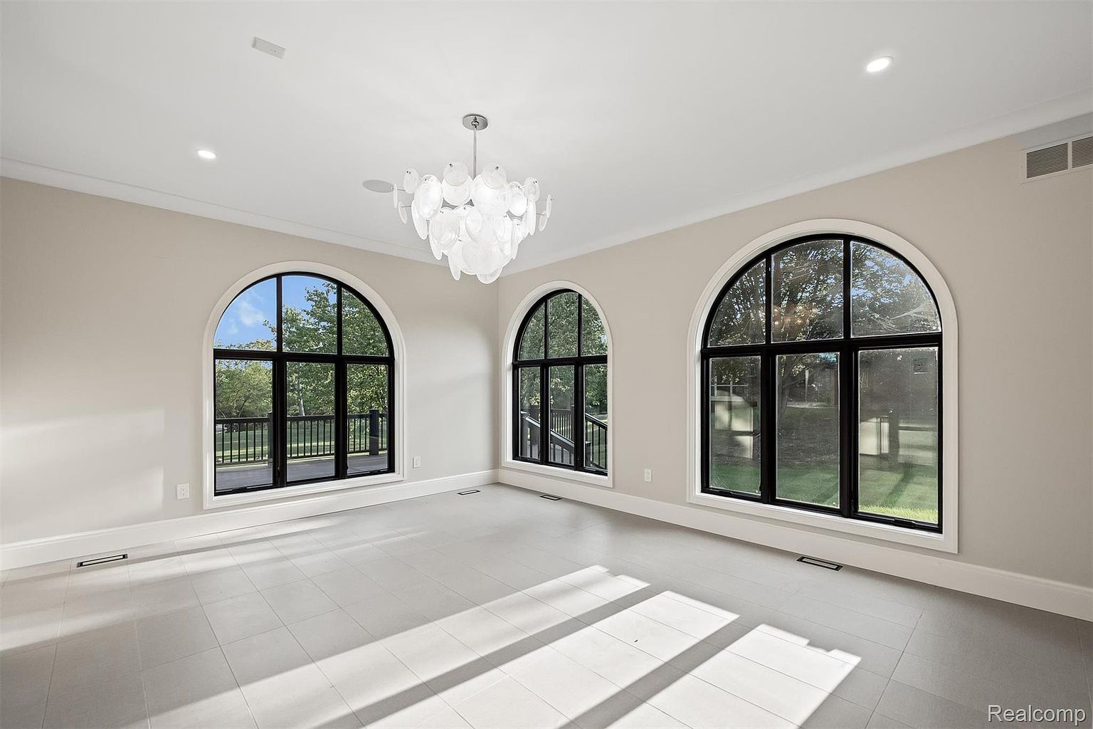 This is a spacious living room featuring three large arched windows that provide ample natural light and views of the outdoors. The room has a neutral color palette with light gray tile flooring and walls, complemented by a modern chandelier. The overall impression is one of elegance and openness, making it an ideal space for relaxation and entertaining.