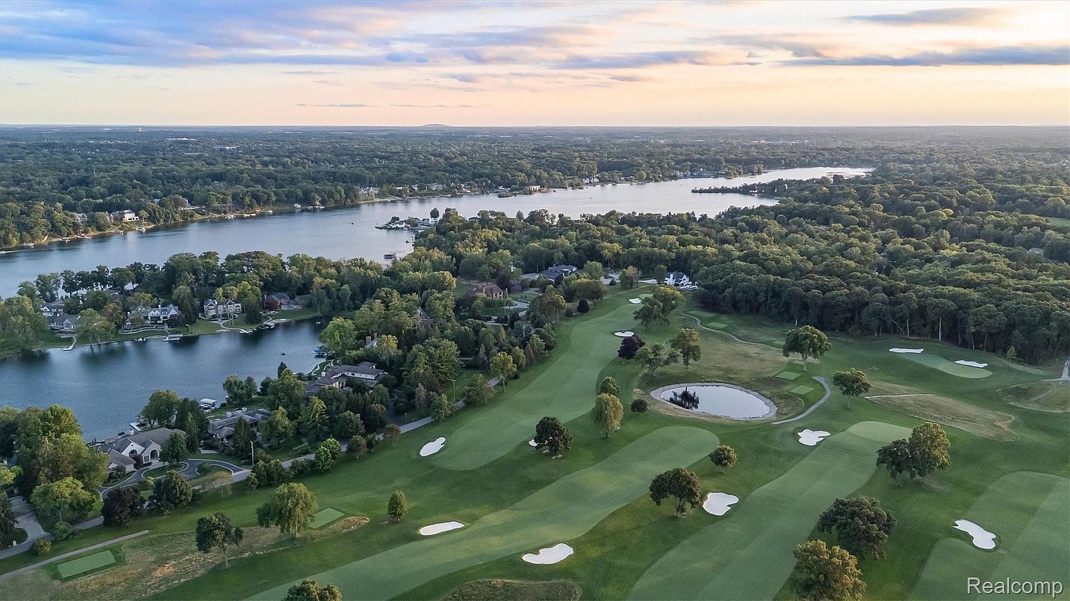 This aerial view showcases a luxurious residential area nestled alongside a serene lake. The landscape features lush greenery, including a well-manicured golf course with sand traps and a small pond. Several upscale homes are visible along the waterfront, blending seamlessly with the natural surroundings under a soft, pastel-colored sky.