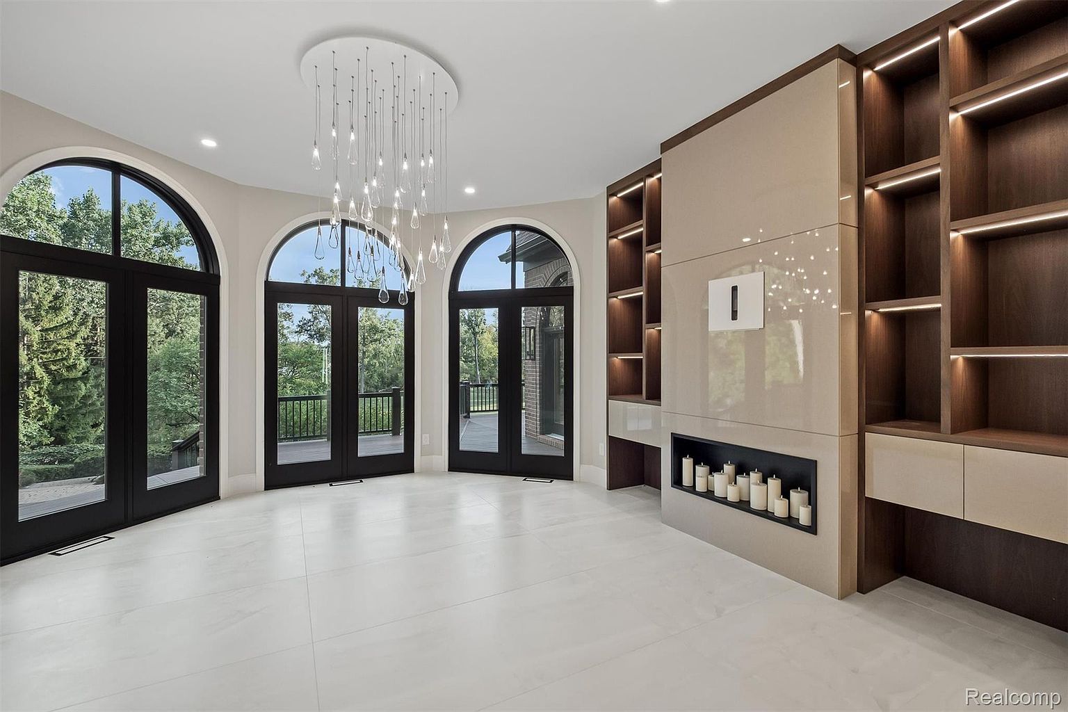 This is a spacious living room featuring three arched doorways/windows with black frames that offer a view of the outdoors. A modern chandelier hangs from the ceiling, and a built-in unit with a fireplace and shelving adds a touch of elegance. The white tile flooring and neutral color palette create a bright and airy atmosphere.