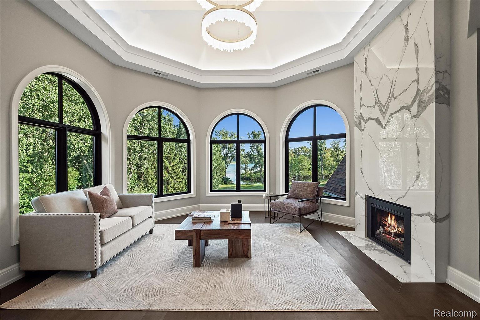 This is an interior shot of a luxurious living room featuring a neutral color palette, large arched windows offering views of lush greenery, and a striking marble fireplace. The room is furnished with a comfortable sofa, an accent chair, and a modern coffee table, all arranged on a patterned area rug. The perspective is from a medium distance, capturing the entire living space and highlighting its elegant design and natural light.