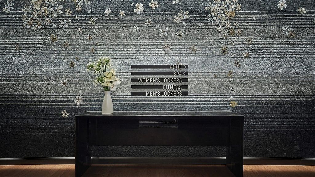 The image showcases an interior space, possibly a hallway or entryway, featuring a dark, glossy console table against a textured wall with floral patterns. A vase of white flowers sits atop the table, adding a touch of elegance. A sign with directions to amenities such as the pool, spa, and fitness center is visible, suggesting this space is within a hotel or luxury apartment complex.