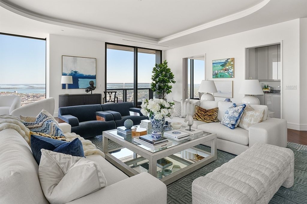This is a luxurious living room featuring a combination of white and navy blue sofas, creating a sophisticated and inviting atmosphere. A mirrored coffee table sits atop a patterned rug, reflecting light and adding to the room's spacious feel. Large windows offer stunning city views, enhancing the upscale ambiance of the space.