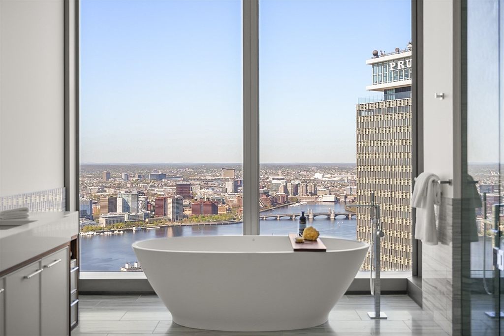 This is a luxurious primary bathroom featuring a freestanding soaking tub positioned in front of a large window with a stunning city view. The bathroom includes modern fixtures, sleek cabinetry, and a glass-enclosed shower, creating a spa-like atmosphere with an emphasis on relaxation and high-end living.