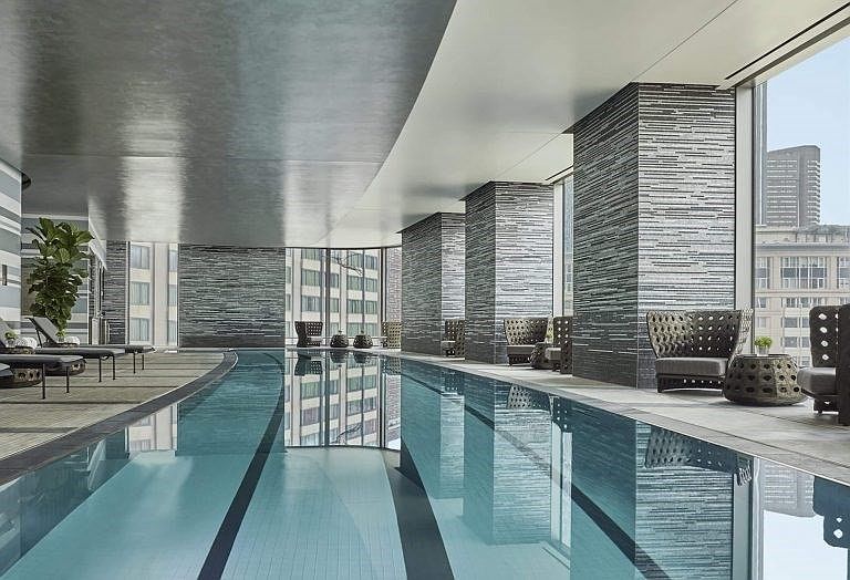 This image showcases a luxurious indoor pool area, featuring a long, sleek pool with clear blue water. The space is surrounded by modern architectural elements, including tiled columns and large windows offering city views. Lounge chairs and stylish seating areas are arranged along the poolside, creating a relaxing and upscale atmosphere.