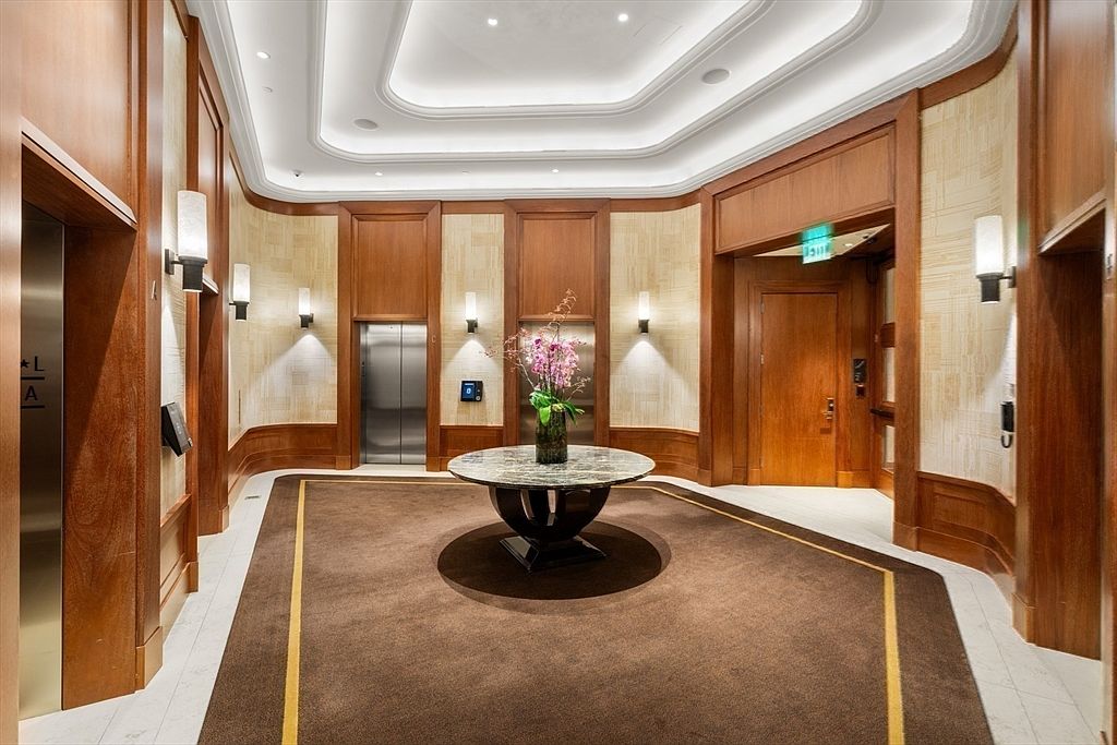 This is an interior shot of a luxurious hallway or lobby area. The space features wood paneling, decorative lighting fixtures, and a round table with a floral arrangement in the center. The flooring is a combination of carpet and tile, and there are elevator doors visible, suggesting a high-end residential building or hotel.