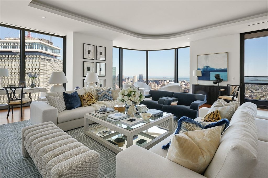 This is a luxurious living room featuring two white sofas adorned with decorative pillows, a navy blue sofa, and a glass-topped coffee table. Large windows offer panoramic city views, enhancing the room's bright and airy atmosphere. The space is elegantly furnished with modern art and stylish decor, creating a sophisticated and inviting ambiance.