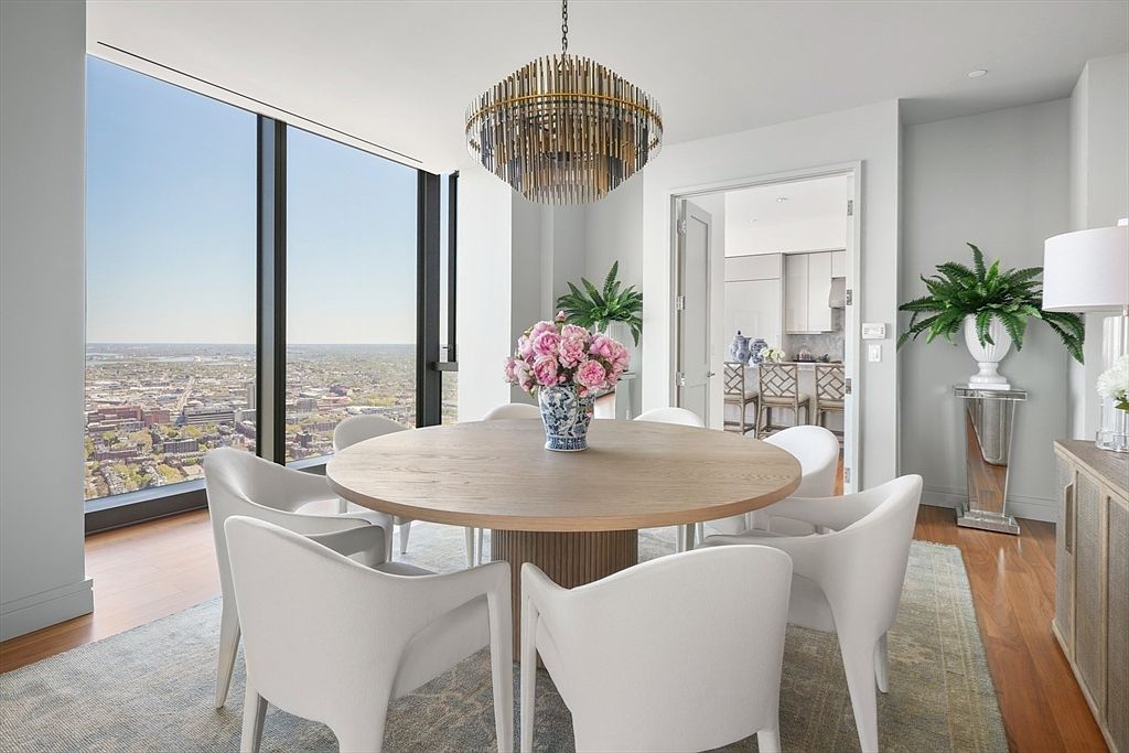 This is an interior shot of a modern dining room featuring a round wooden table surrounded by white chairs. A striking chandelier hangs above the table, and a large window offers a panoramic city view. The room is decorated with plants and flowers, creating a bright and inviting atmosphere.