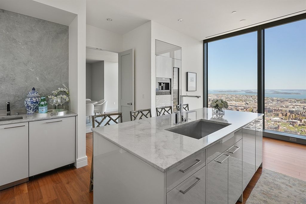 This is a bright and modern kitchen with sleek white cabinetry and a large island featuring a marble countertop and stainless steel sink. Floor-to-ceiling windows offer stunning city views, enhancing the luxurious feel of the space. The kitchen also includes built-in appliances and a stylish bar area, perfect for entertaining.