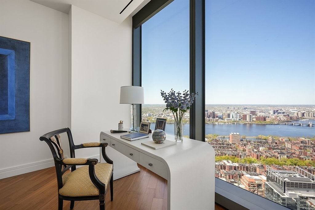 This interior shot showcases a modern office space with a stunning city view. A sleek, white desk is positioned in front of a large window, complemented by a stylish chair. The room features hardwood flooring, a blue painting on the wall, and a minimalist design, creating a sophisticated and productive atmosphere.