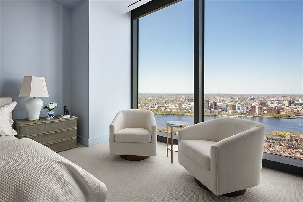 This primary bedroom features a serene and luxurious atmosphere with a stunning city view through large windows. Two comfortable armchairs and a small side table create a cozy seating area by the window. The room is decorated in a calming color palette, complemented by elegant furnishings such as a bedside table with a lamp and a neatly made bed.