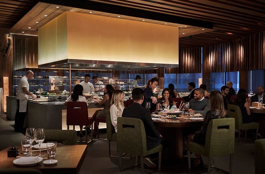 This is an interior shot of a modern dining room in a restaurant setting. The room features a large open kitchen area where chefs are preparing food, with a dining area in the foreground where patrons are seated at tables. The space is illuminated by warm lighting, creating an inviting and upscale atmosphere, perfect for showcasing a high-end dining experience.