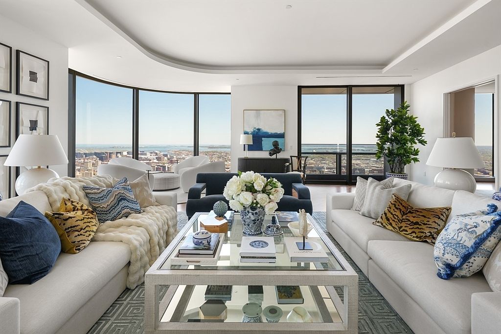 This is a bright and luxurious living room featuring two white sofas adorned with decorative pillows, a glass-topped coffee table with decorative items, and large windows offering a panoramic city view. The room is decorated in a modern style with a neutral color palette, creating an elegant and inviting atmosphere. The perspective is from the center of the room, showcasing the symmetry and spaciousness.