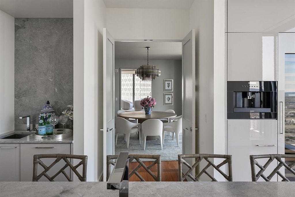 This image showcases a dining area viewed through an open doorway, featuring a round wooden table surrounded by white chairs. A modern chandelier hangs above the table, and artwork adorns the walls. The space is bright and airy, with a sophisticated and inviting atmosphere, complemented by a glimpse of a bar area to the left and a built-in coffee machine to the right.