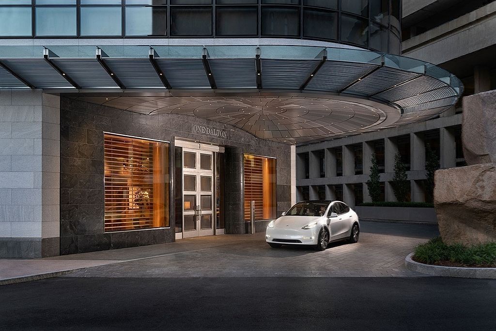 The image showcases the grand entryway of "One Dalton", featuring a modern architectural design with a curved canopy and stone facade. A white Tesla is parked in front, adding a touch of luxury. The overall impression is sophisticated and inviting, highlighting the building's upscale appeal.