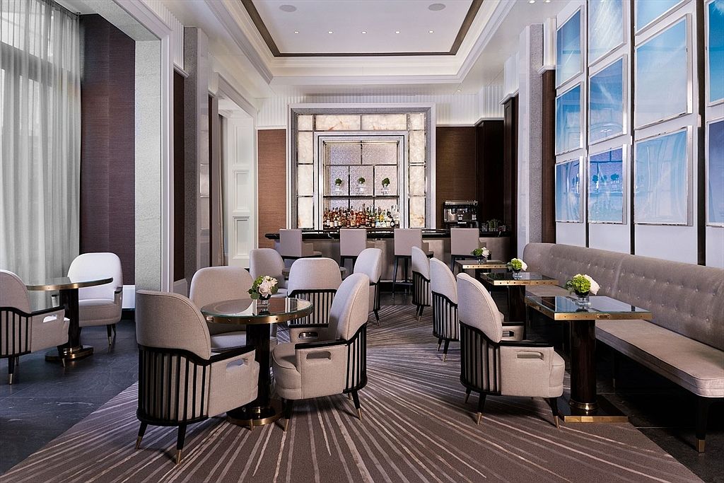 This is an elegant dining area featuring comfortable armchairs around small, round tables, and a long cushioned bench along the wall. A bar area is visible in the background, suggesting a sophisticated lounge or restaurant setting. The room is decorated in neutral tones with dark wood accents, creating a luxurious and inviting atmosphere.