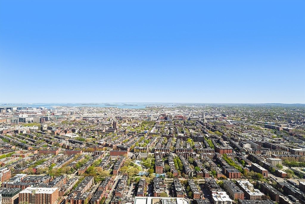 This aerial view showcases a sprawling cityscape under a clear blue sky, highlighting the density of residential and commercial buildings interspersed with green spaces. The perspective offers a comprehensive look at the urban layout, emphasizing the scale and interconnectedness of the neighborhood.