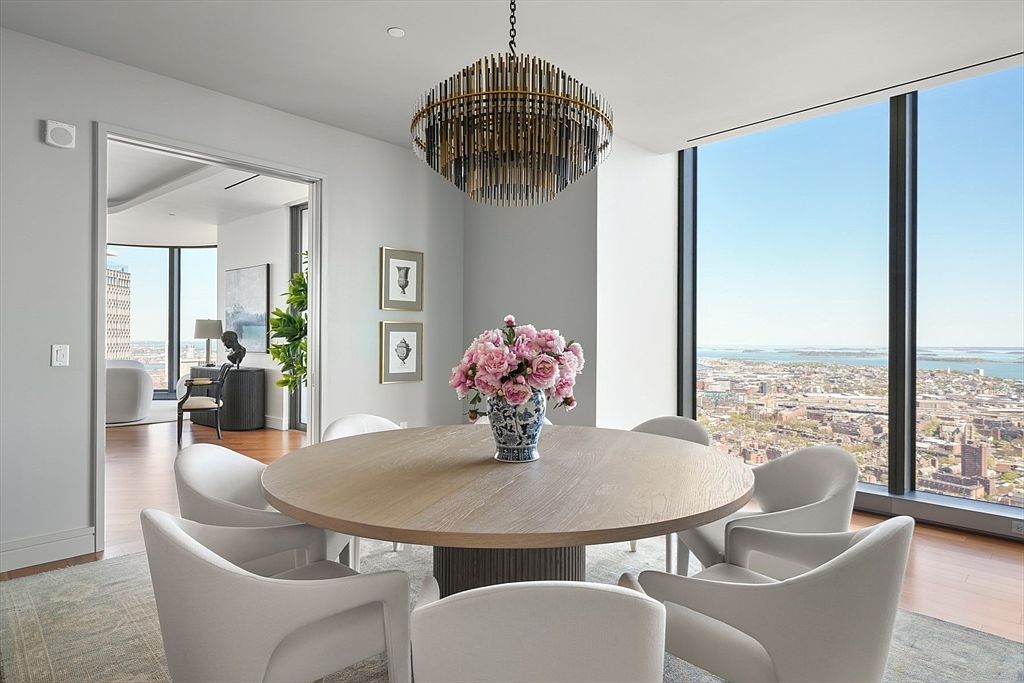 This is an interior shot of a modern dining room featuring a round wooden table with a floral centerpiece and surrounded by six white chairs. A striking chandelier hangs above the table, and a large window offers a panoramic city view. The room exudes elegance and sophistication, perfect for upscale dining and entertaining.