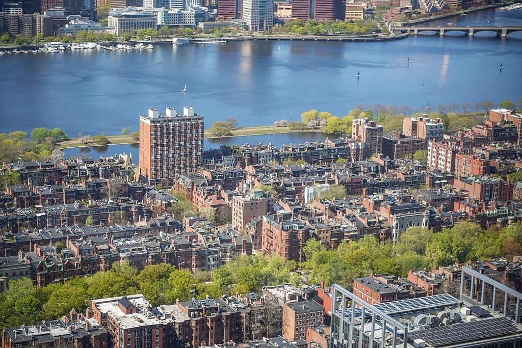 This aerial view showcases a dense urban landscape with closely packed brick buildings and lush green trees interspersed throughout. A wide river dominates the background, reflecting sunlight and featuring a bridge in the distance. The scene provides a sense of established community and proximity to waterfront amenities.