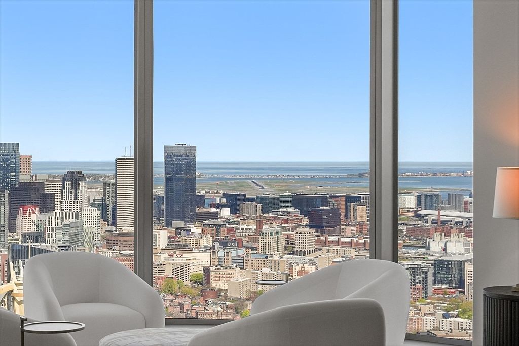 This interior shot showcases a luxurious living room with floor-to-ceiling windows offering a panoramic city view. Two modern, white armchairs and an ottoman are arranged to maximize the view, creating a sophisticated and inviting space. The room's design emphasizes natural light and urban elegance, making it a desirable feature for potential buyers.