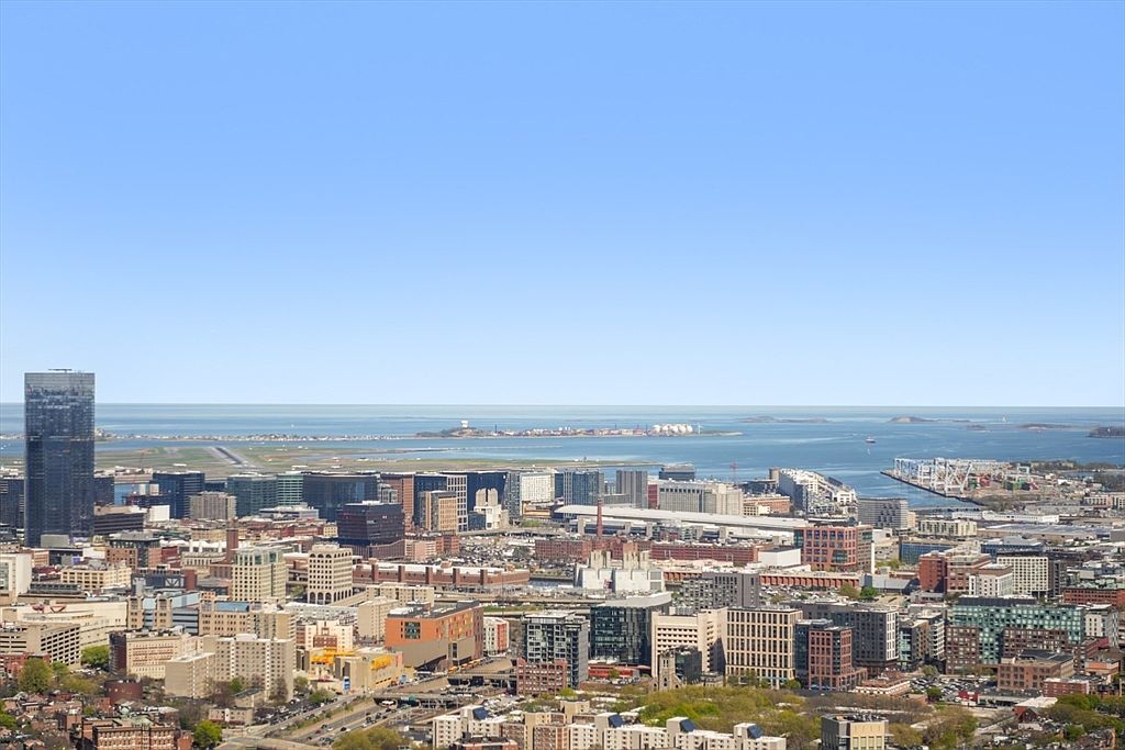 This aerial view showcases a sprawling cityscape with a prominent skyscraper on the left. The city extends towards a body of water, where islands and distant landmasses are visible under a clear blue sky. The image provides a sense of the city's scale and its proximity to the coast, highlighting its urban density and scenic location.